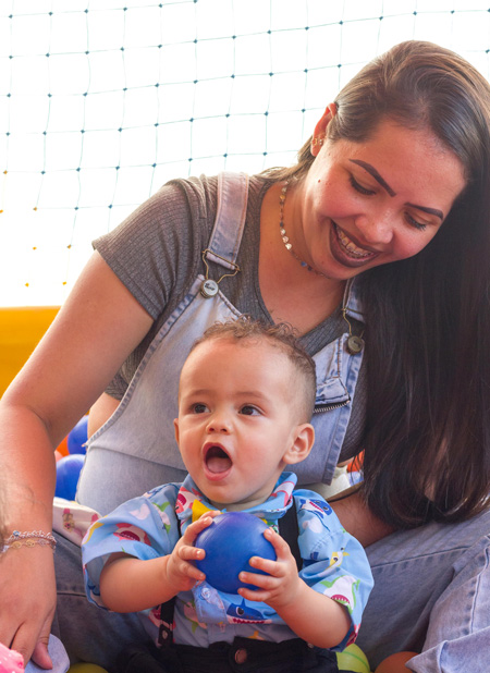 eine Mutter mit langen dunklen Haaren sitz mit ihrem Kind auf dem Schoss und lächelt, das Kind hält in seinen Händen einen Ball, beide spielen im Familienzentrum Alve