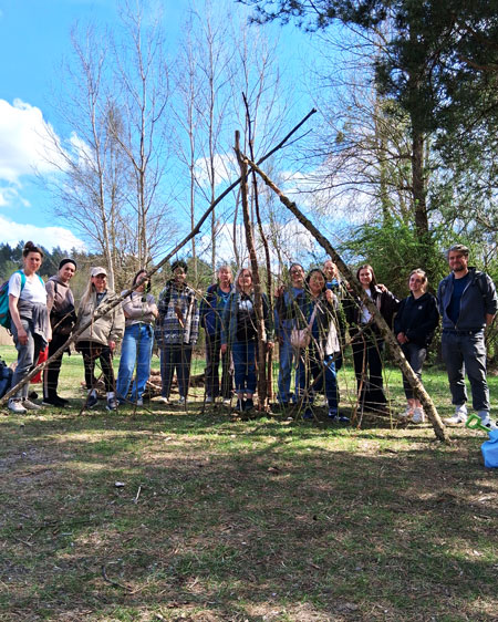 12 Erzieher*innen stehen auf einer Wiese hinter einem Tipi gebaut aus Ästen