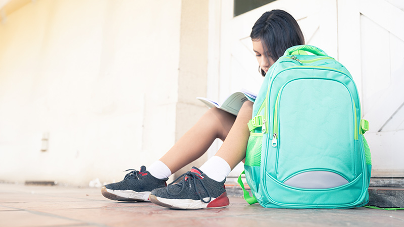 Mädchen sitzt mit grünem Schulranzen auf dem Boden